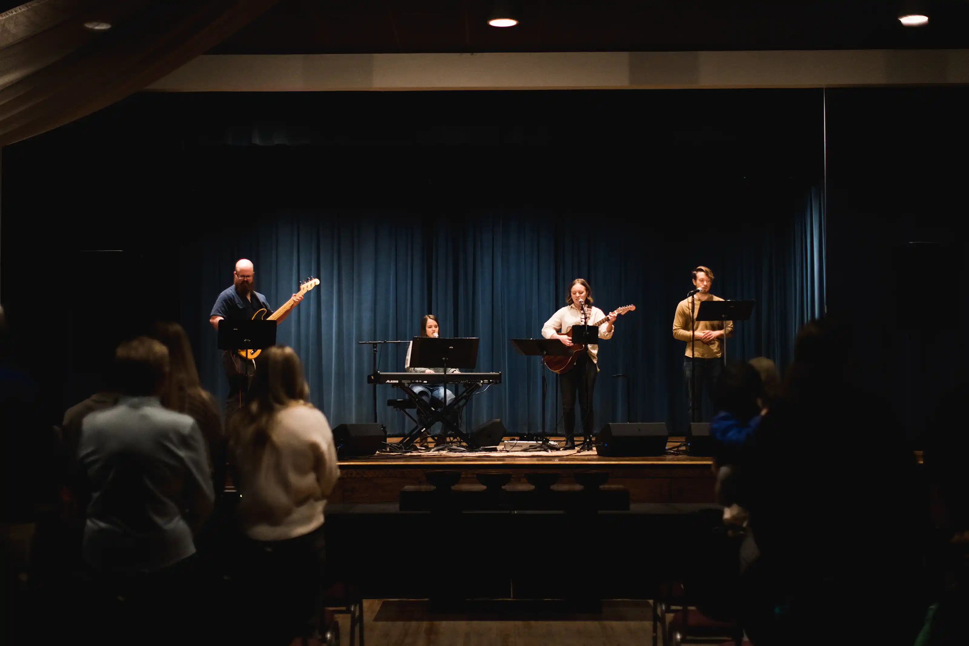 A worship band—bass, piano, guitarist, vocalist—singing on stage in an auditorium