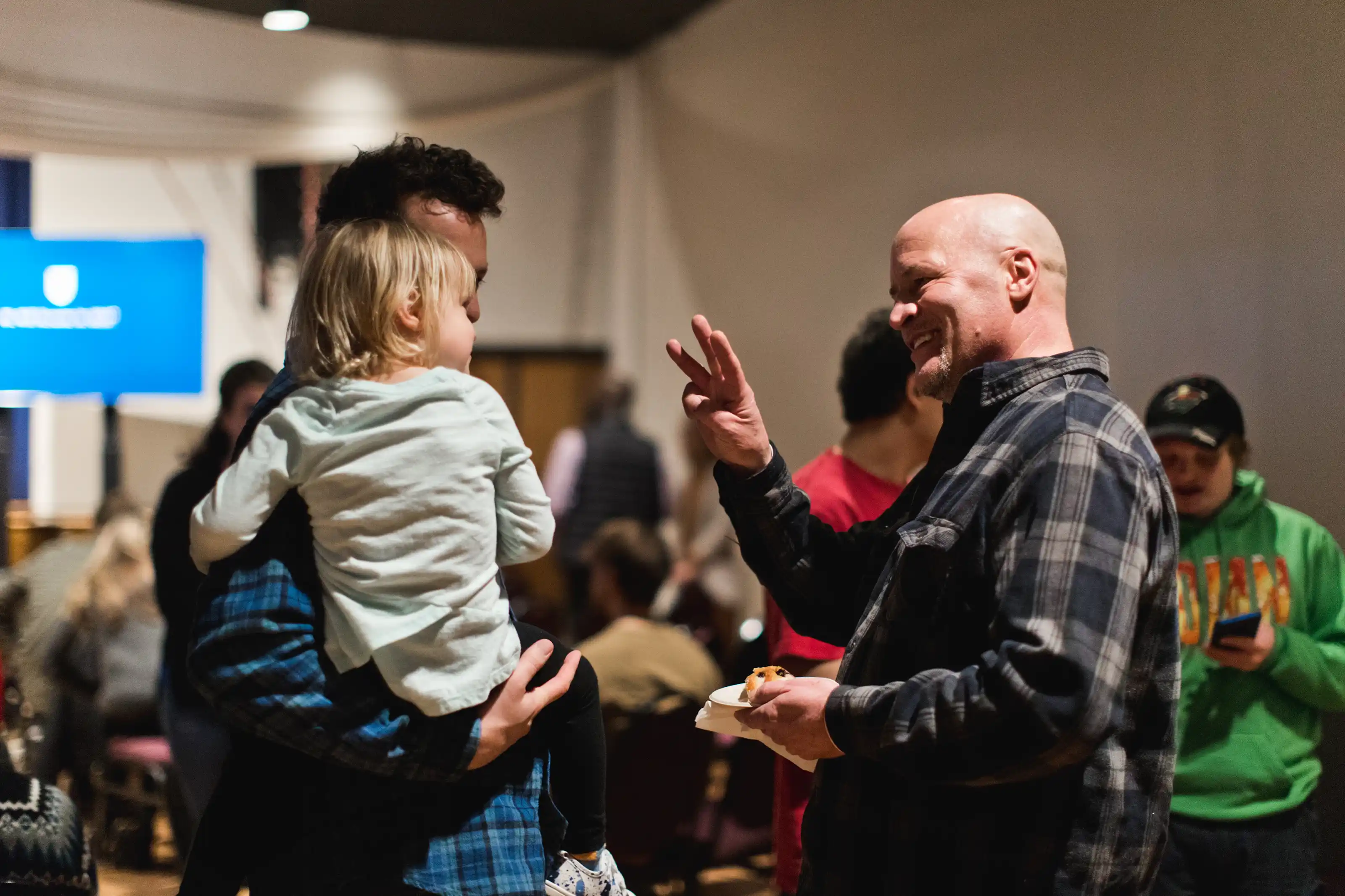 A friendly churchgoer waving at a baby girl being held by her dad in a busy, buzzing church auditorium.
