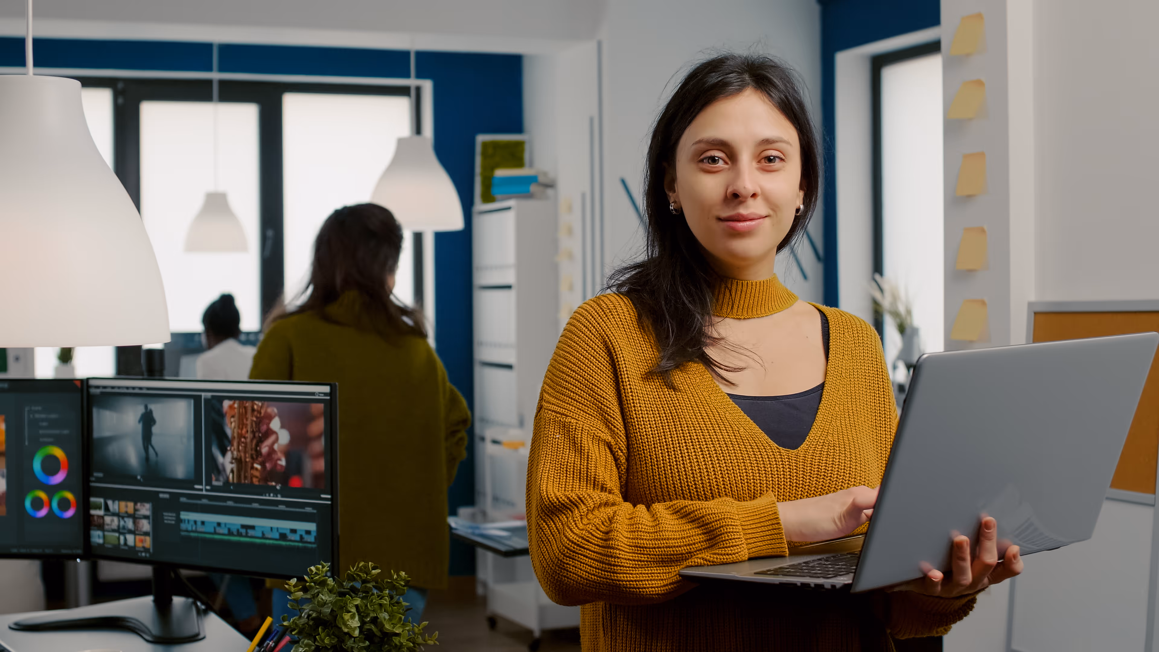 Femme souriante tenant un ordinateur portable dans un bureau moderne avec un poste de travail de montage vidéo en arrière-plan.