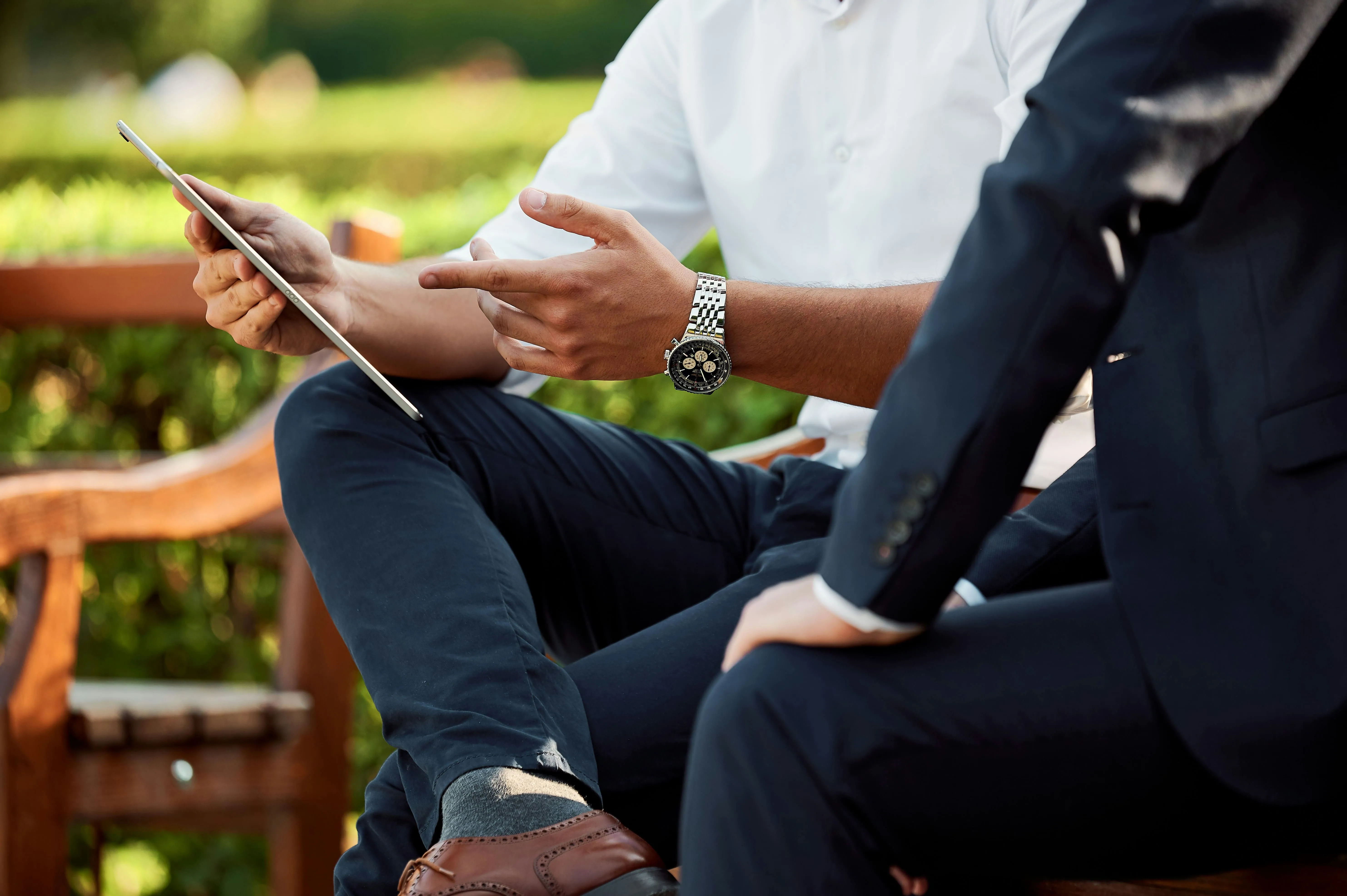 Zwei Fachleute sitzen auf einer Holzbank im Freien und diskutieren etwas, während einer von ihnen ein Tablet hält. Der eine trägt ein weißes Hemd und der andere einen dunklen Anzug.