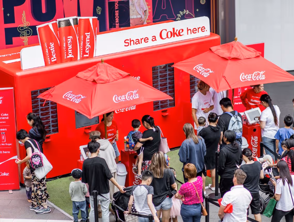 Les gens font la queue devant un stand Coca-Cola avec des parapluies rouges et des distributeurs automatiques de la marque.