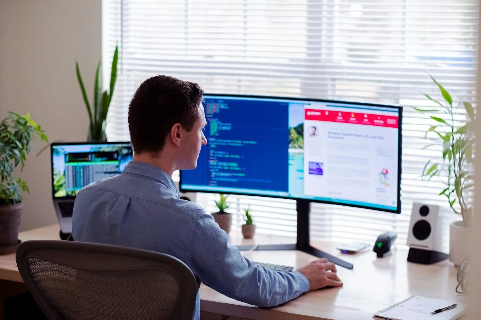 A man working at a home office with multiple monitors, one showing code and the other displaying a website, surrounded by plants and sunlight.