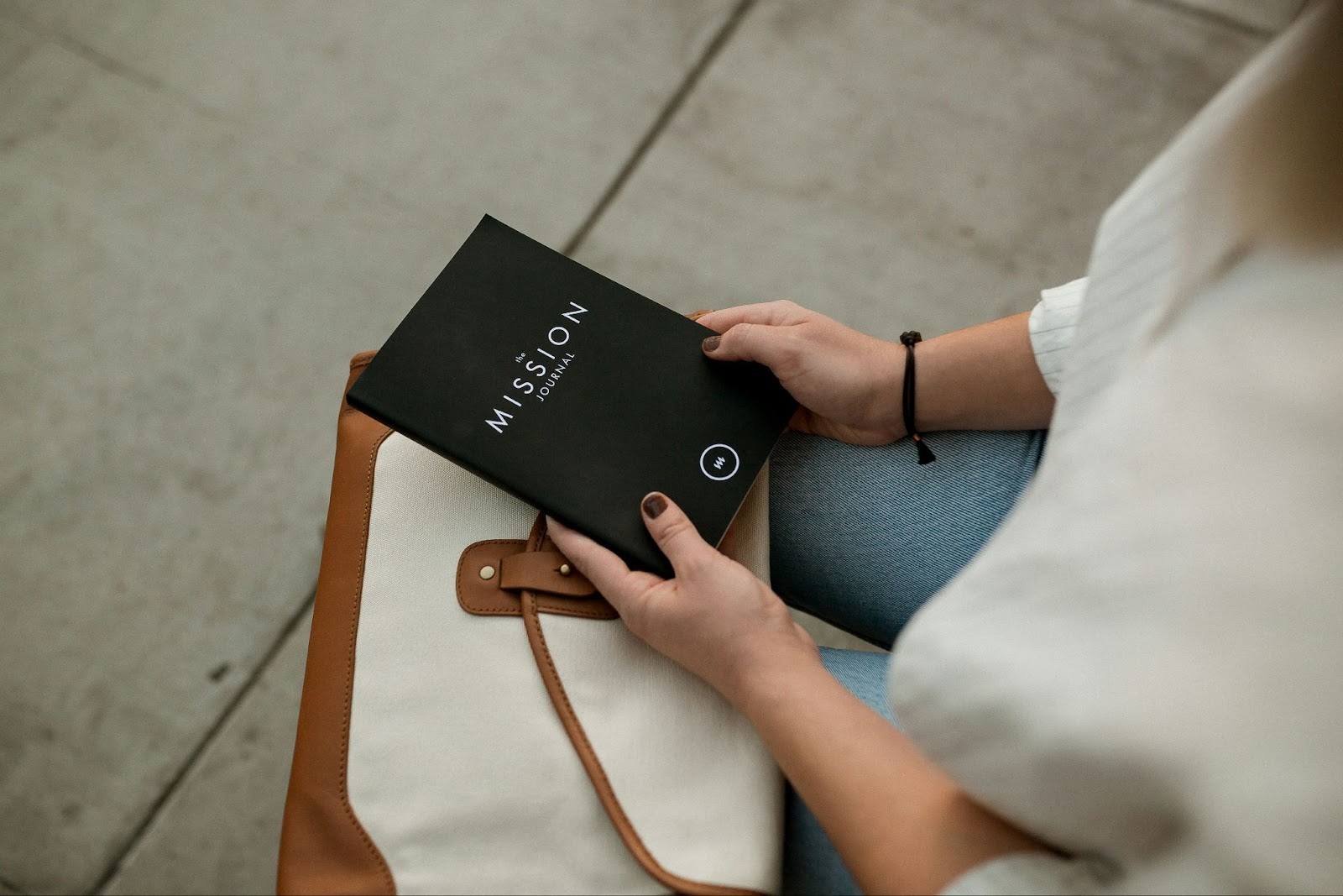 A person holding a black notebook titled “Mission Journal” while sitting on a bench with a beige and brown bag on their lap.