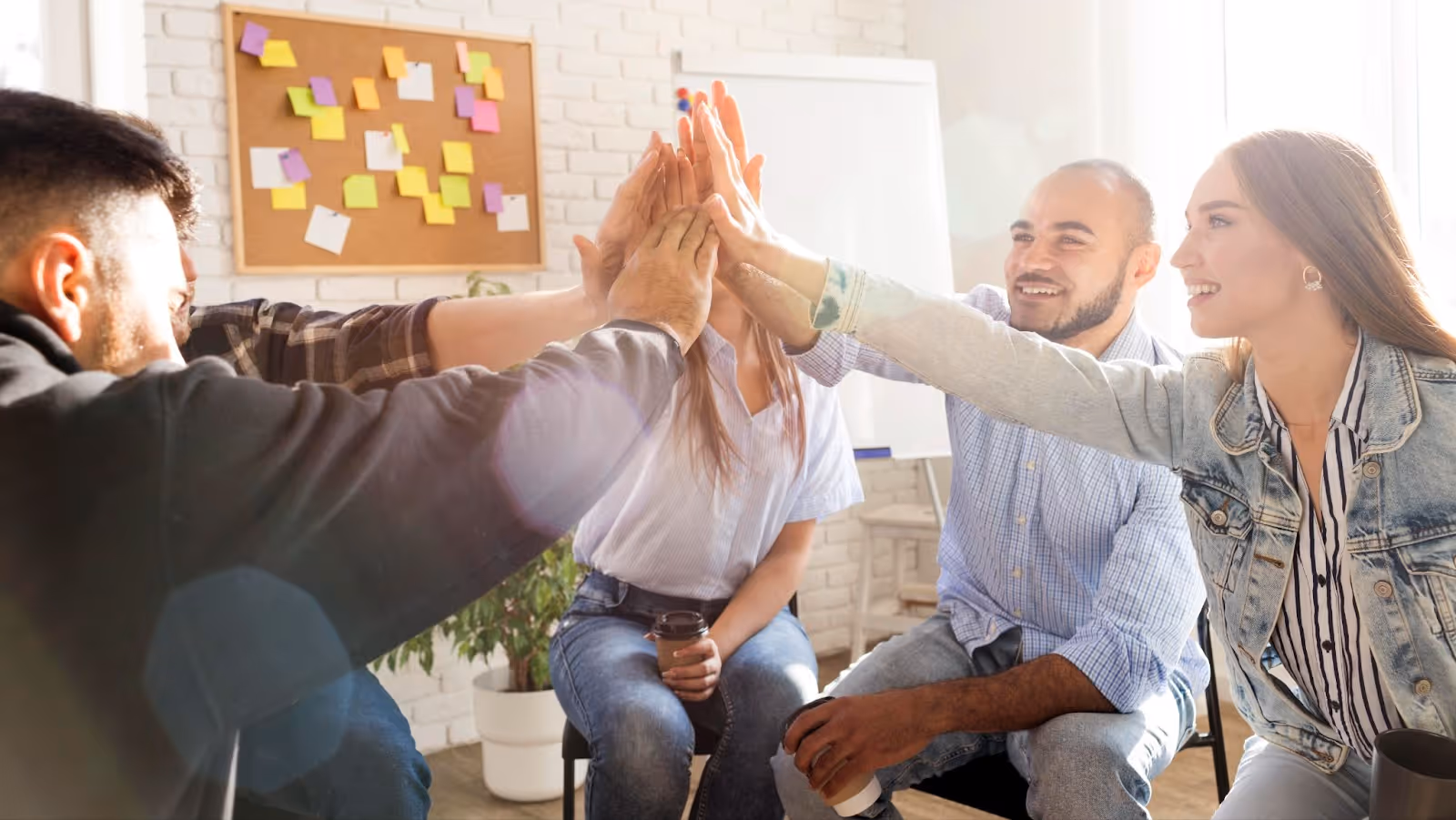 Group of coworkers enthusiastically doing a group high-five in a bright office space with sticky notes on a corkboard in the background.