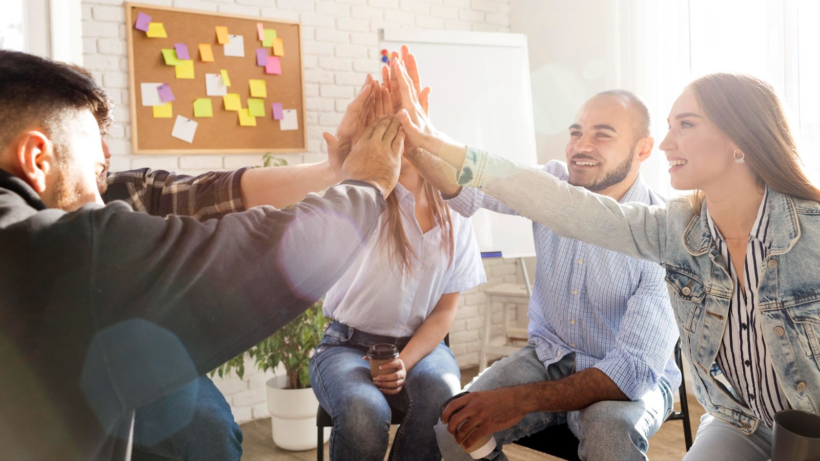 Group of coworkers enthusiastically doing a group high-five in a bright office space with sticky notes on a corkboard in the background.