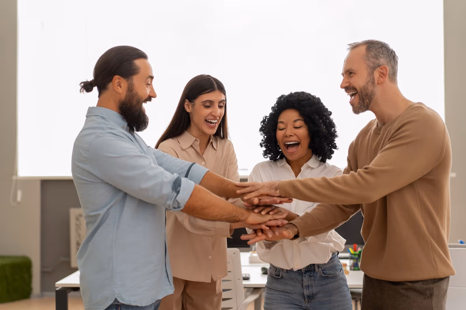 Four colleagues standing in a circle with their hands stacked together, smiling and celebrating teamwork in a modern office.