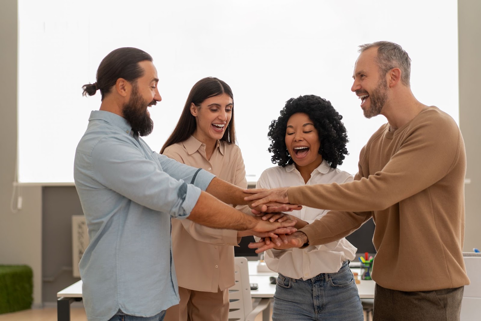 Four colleagues standing in a circle with their hands stacked together, smiling and celebrating teamwork in a modern office.