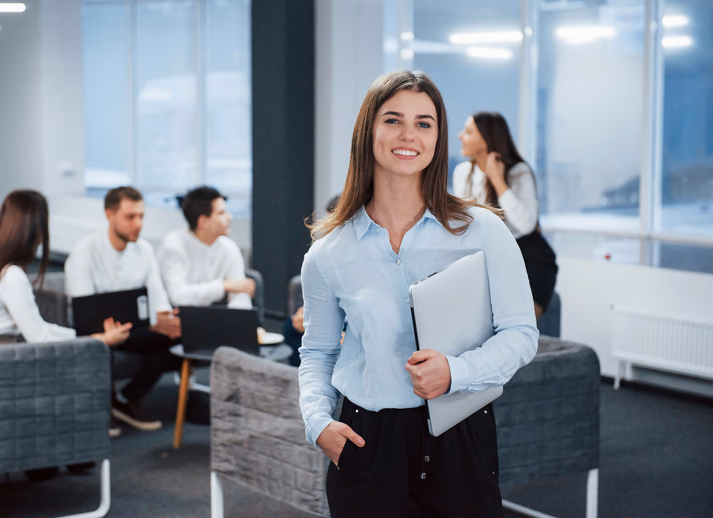 Smiling woman in light blue shirt holding laptop in office with coworkers talking in background.