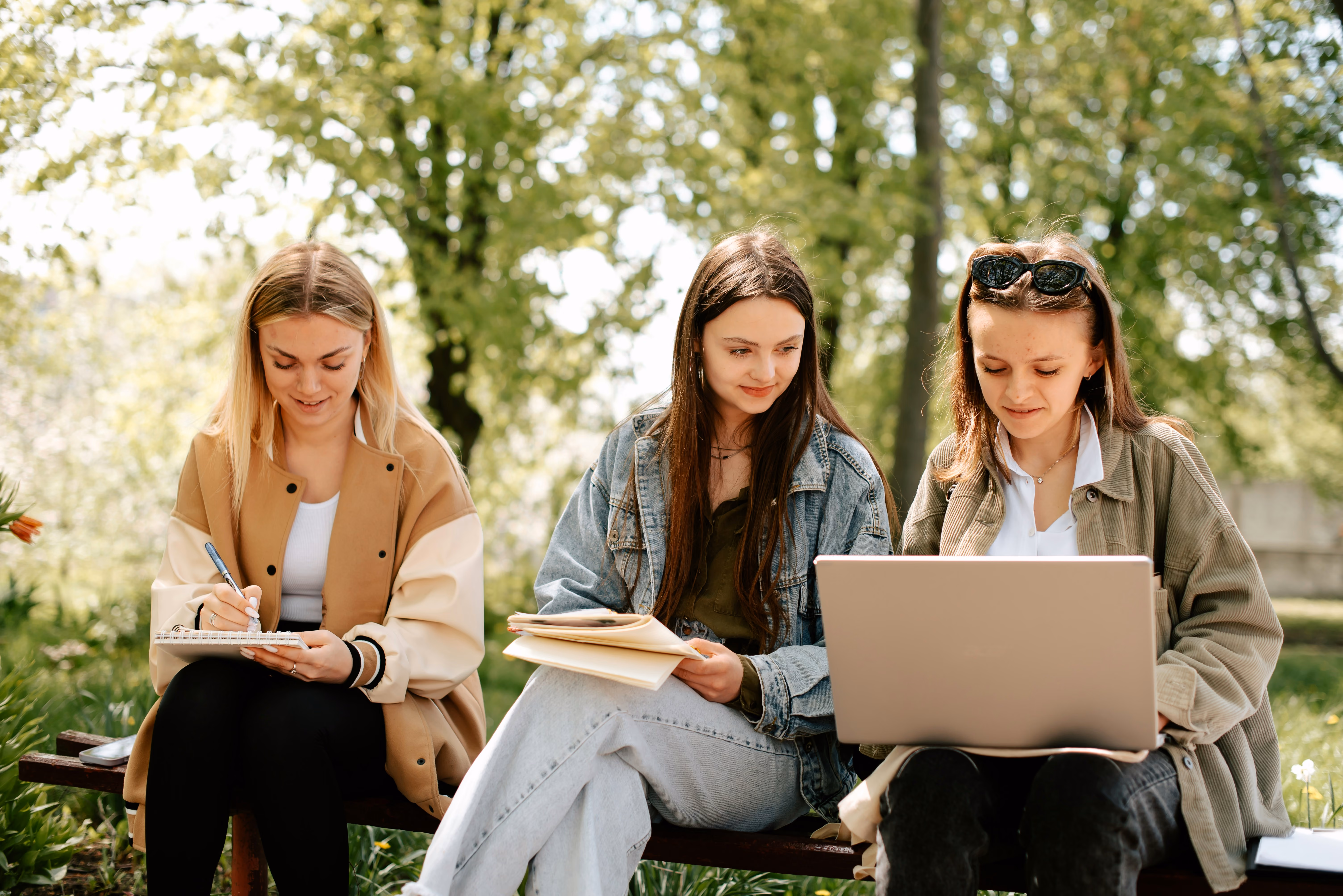 Three young women studying outdoors on a bench with notebooks and a laptop under trees.