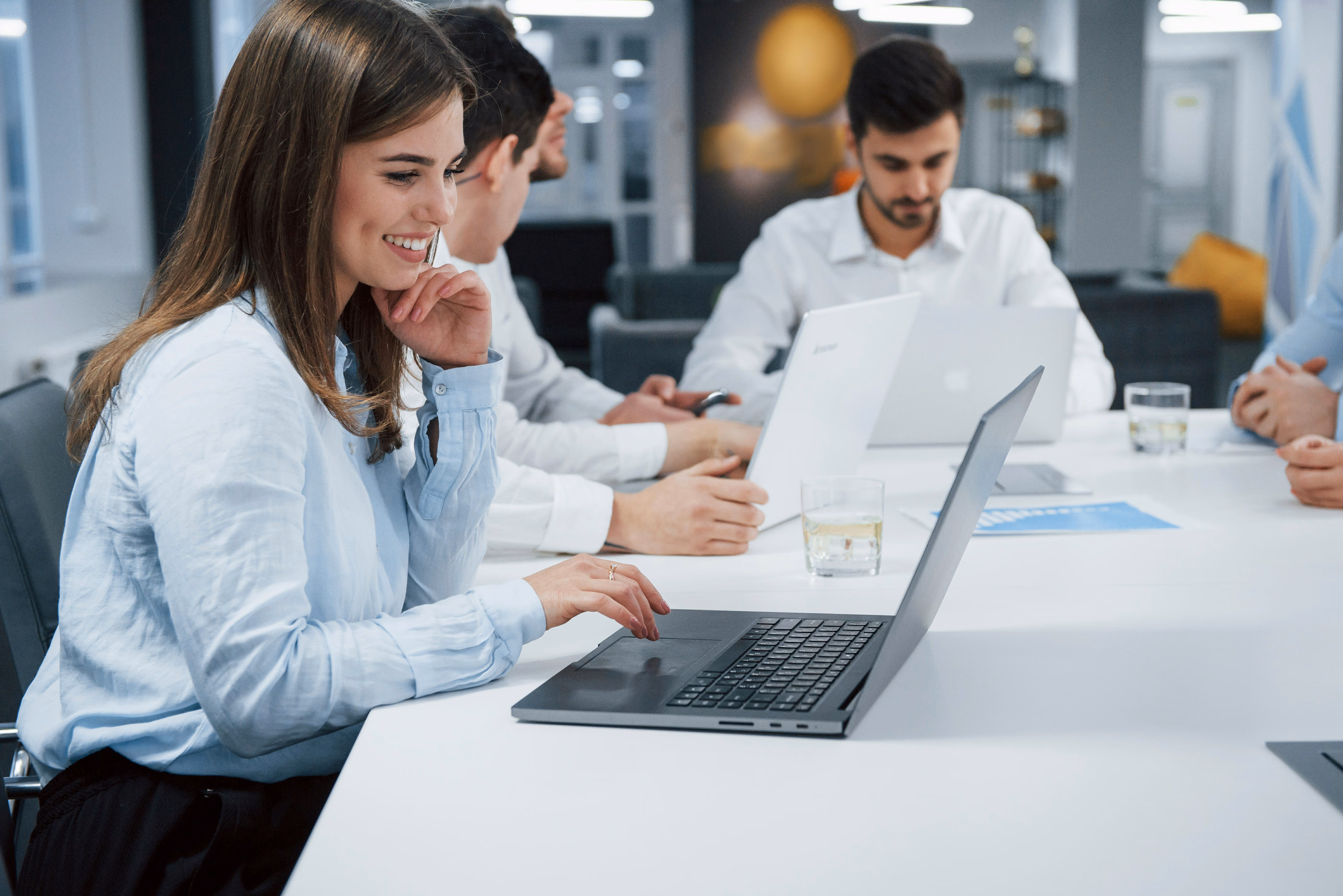 Smiling woman working on a laptop in a meeting with three colleagues using laptops and phones.