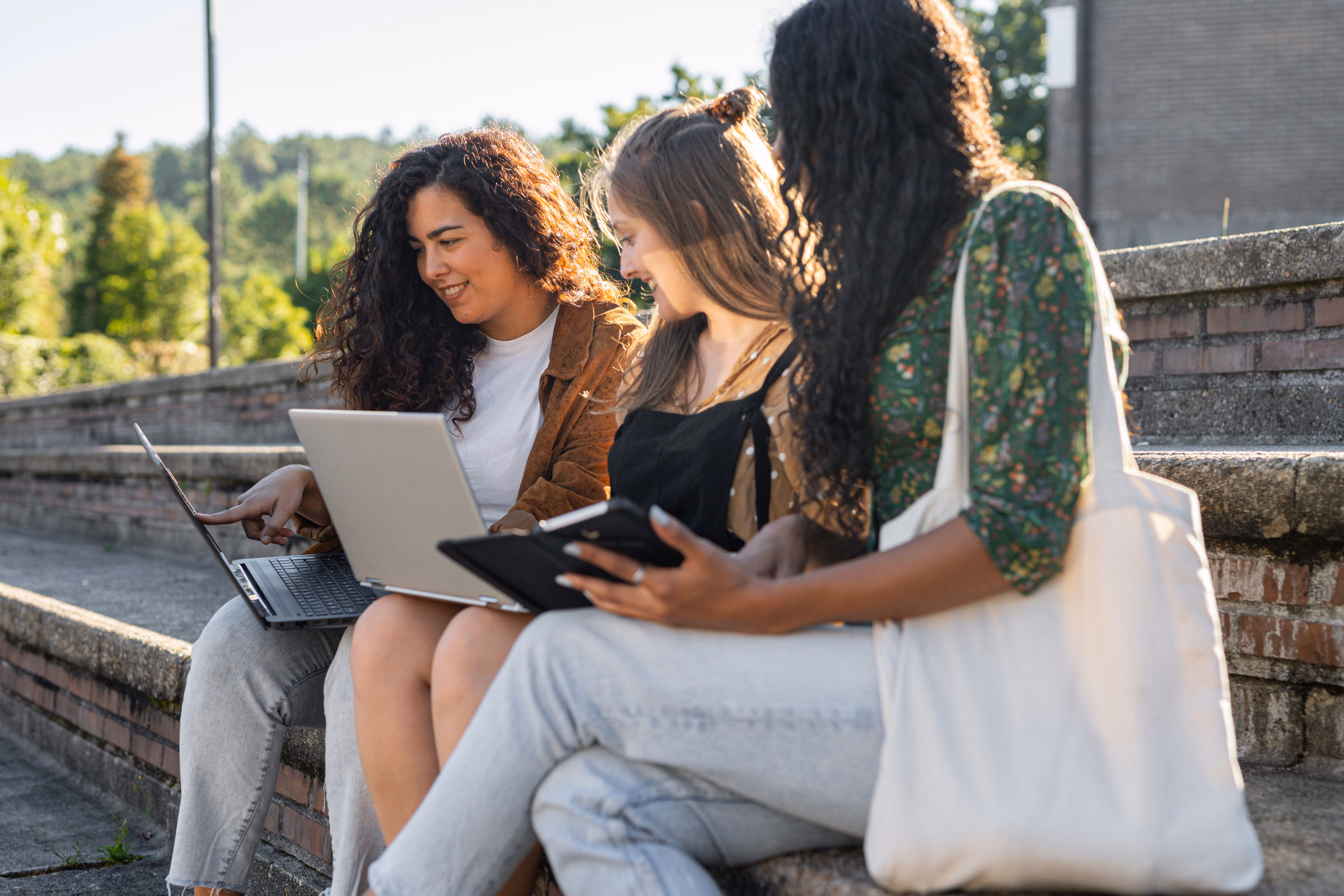 Three young women sitting on outdoor steps sharing a laptop and tablet while chatting.