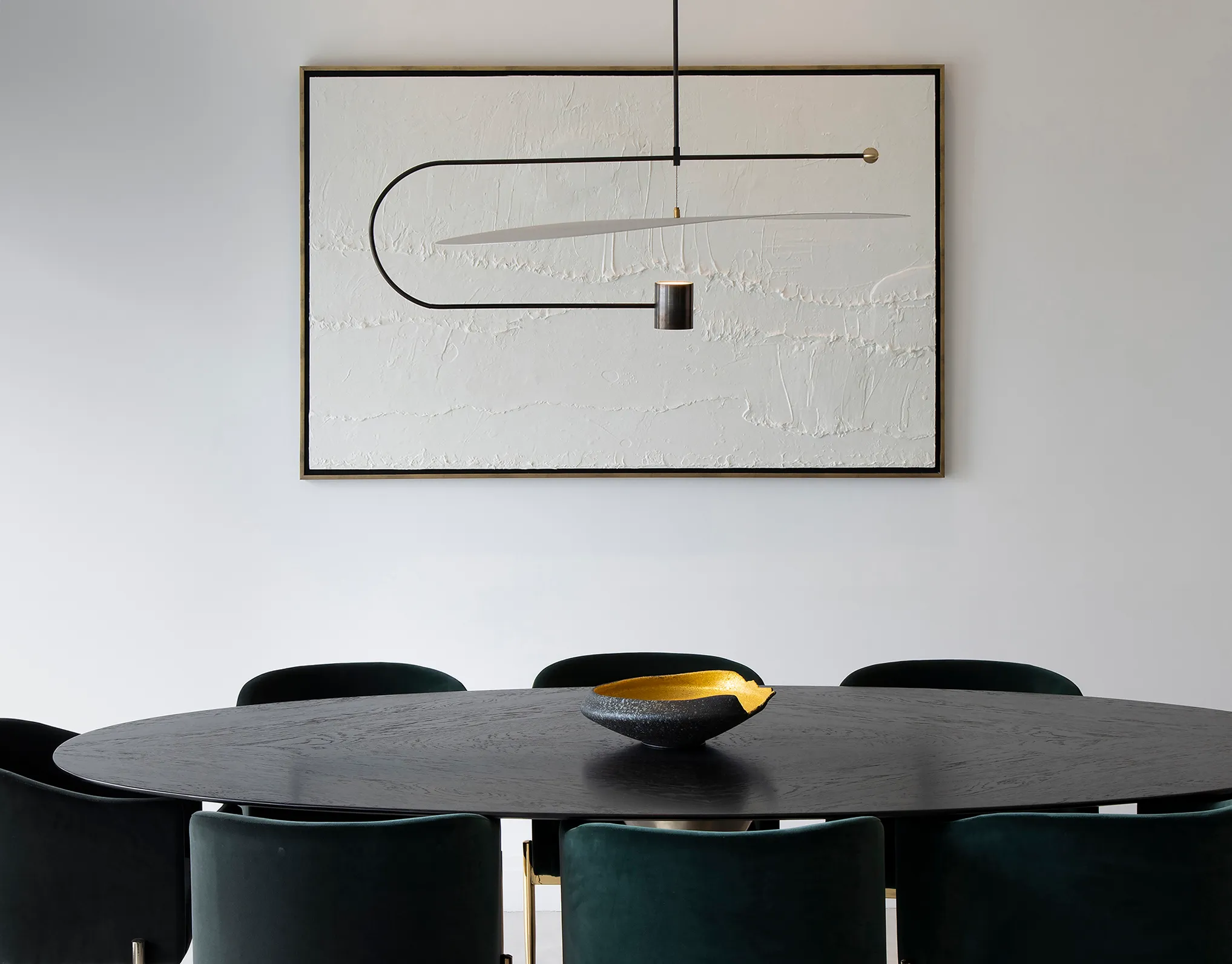 Modern black oval dining table with a decorative bowl, surrounded by dark green chairs and a minimalist pendant light with a black and white abstract art piece on the wall.