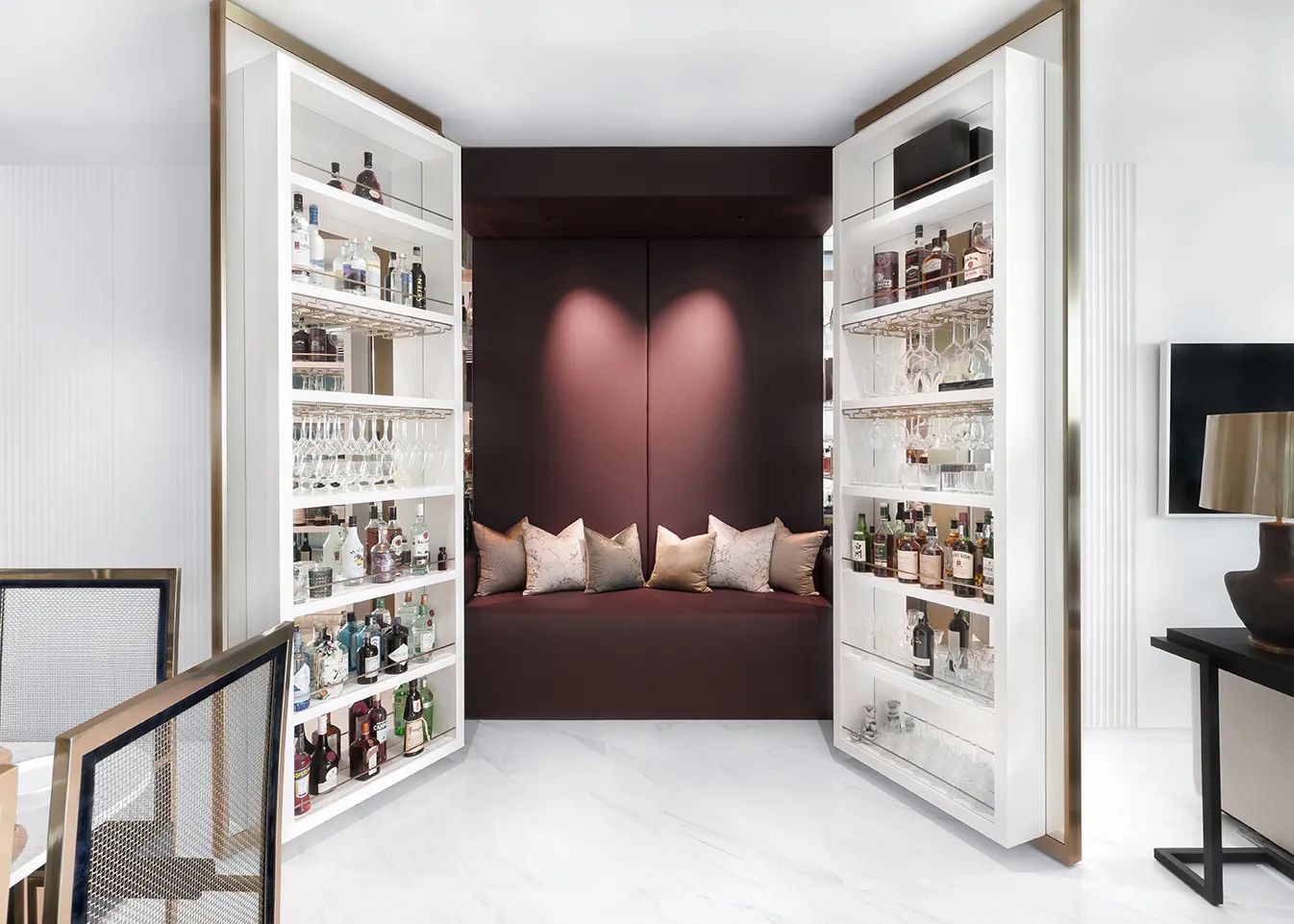 Modern home whisky room with built-in shelves stocked with bottles and glasses, and a burgundy cushioned seating nook with pillows.
