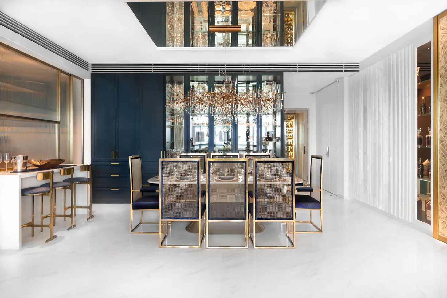 Modern dining room with gold-framed chairs, oval glass table, intricate chandelier, and blue cabinetry with mirrored panels.