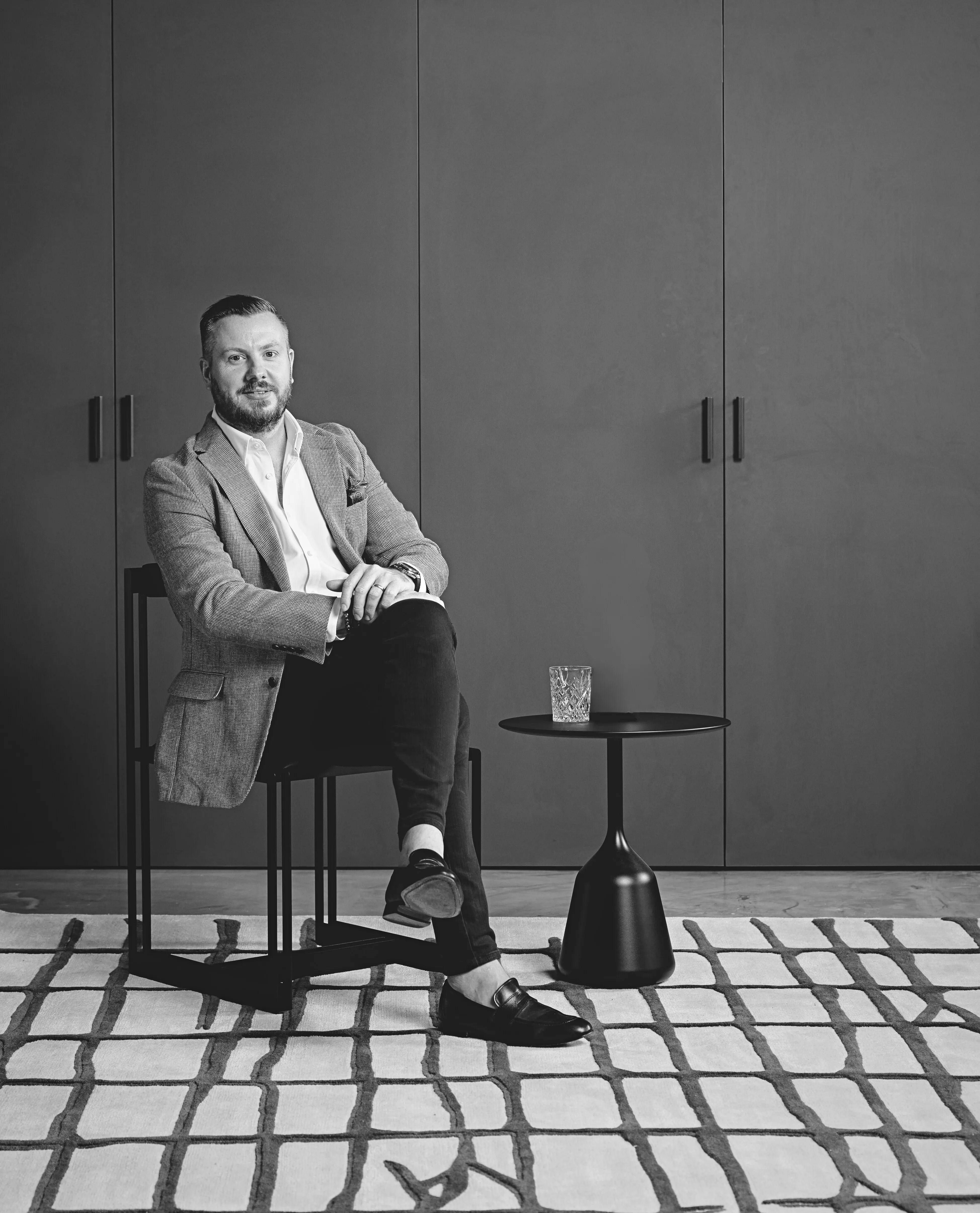 Man in a blazer and loafers sitting cross-legged on a modern chair next to a small round table with a glass on it.