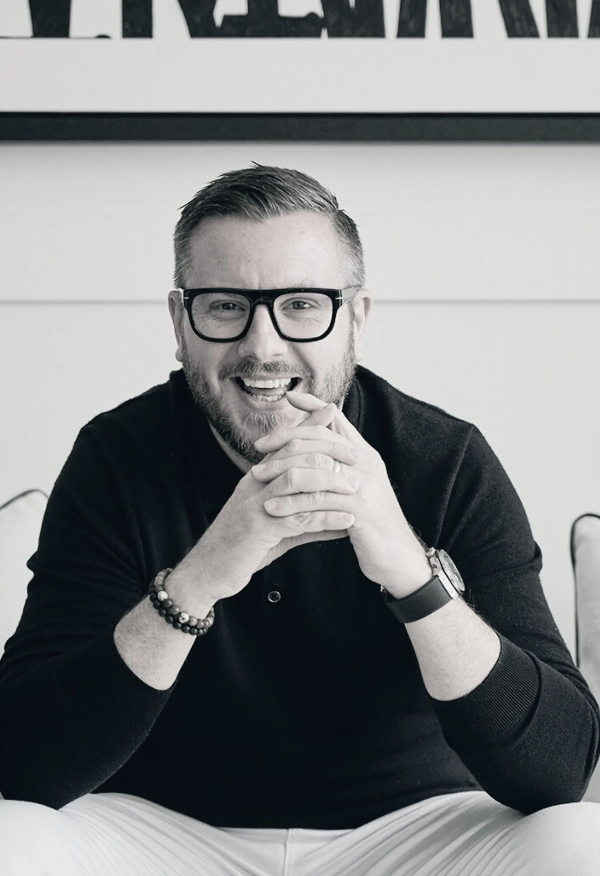 Smiling man with glasses and a beard sitting on a couch with hands clasped.