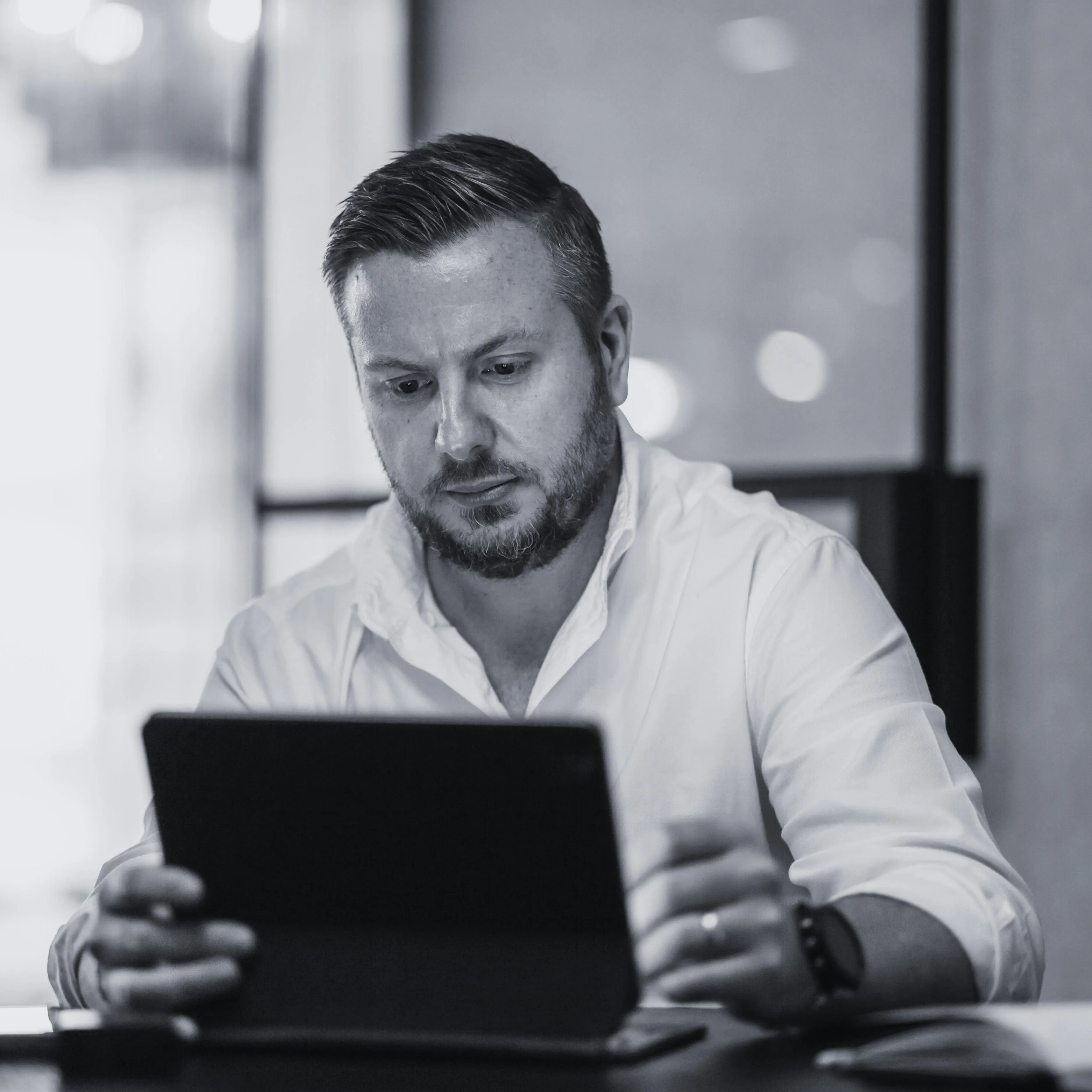Man with short hair and beard wearing a white shirt, focused on using a tablet at a desk.