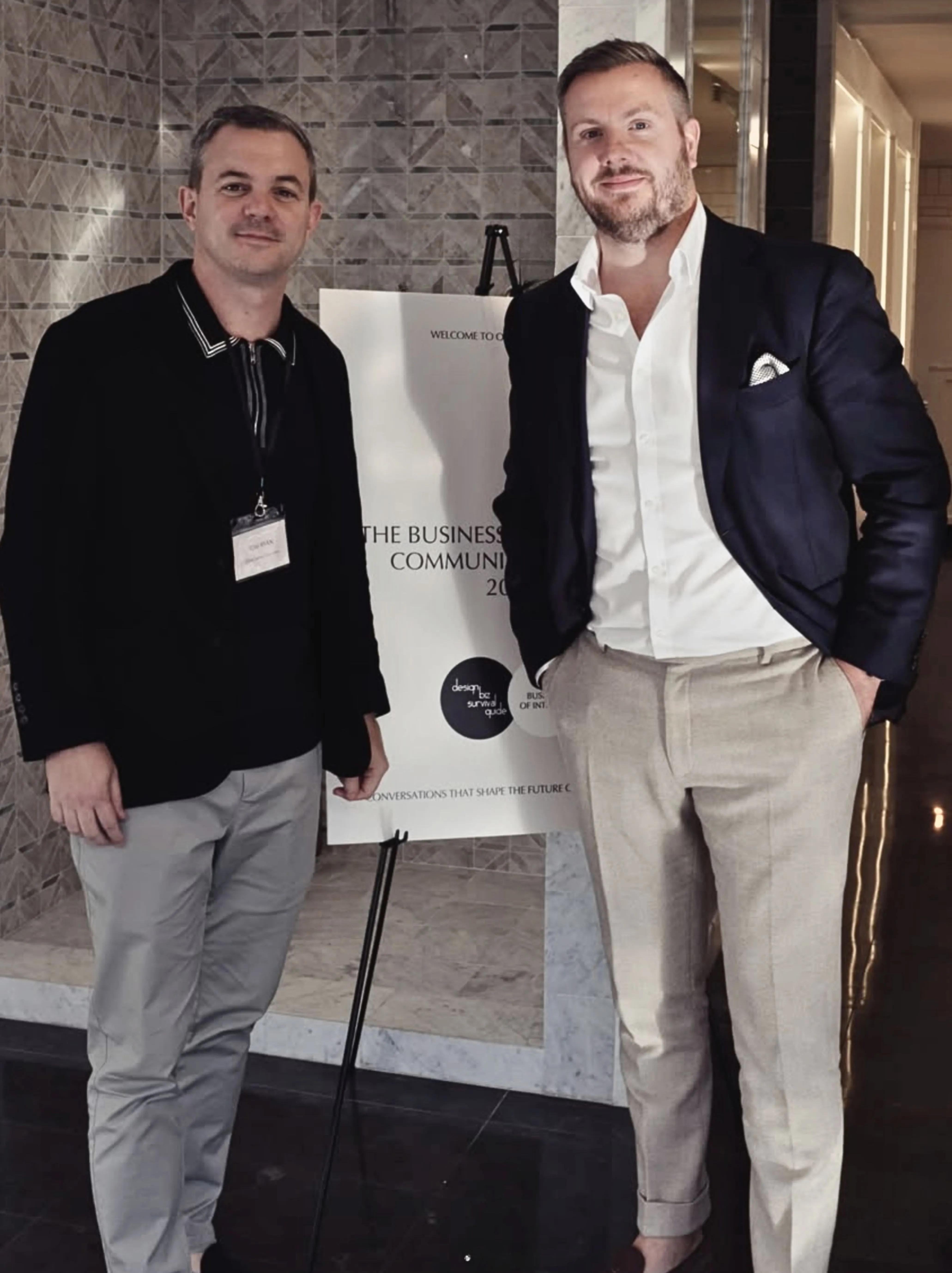 Two men standing indoors near a sign on an easel in a tiled lobby, dressed in business casual attire.