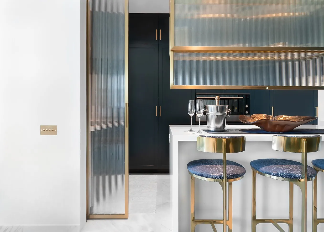 Modern kitchen with blue cushioned gold stools, white island, champagne bucket, and dark cabinetry.