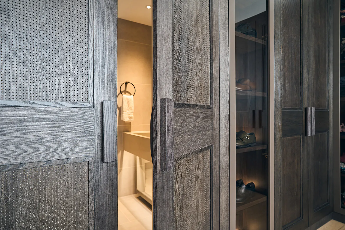 Wooden sliding doors partially open revealing a bathroom with towel on ring and a closet with shoes.
