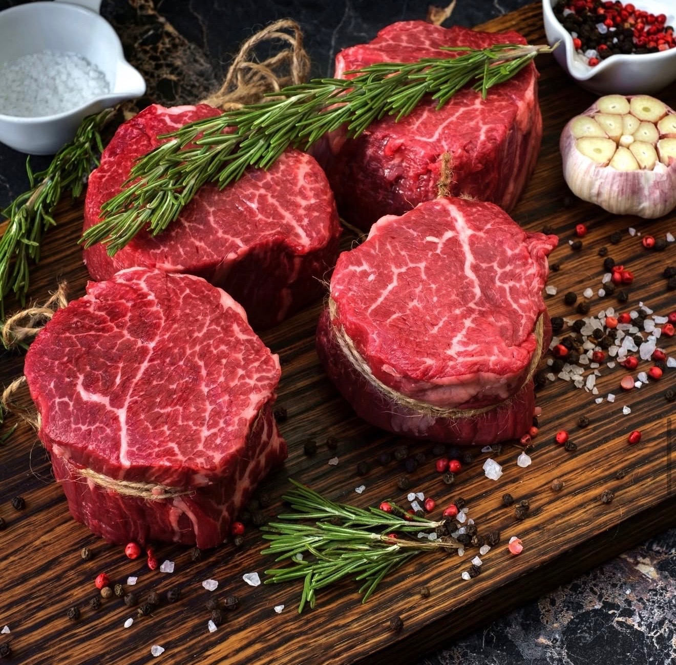 Fresh halal beef filet cuts tied with twine and garnished with rosemary, garlic, coarse salt, and peppercorns on a rustic wooden board.