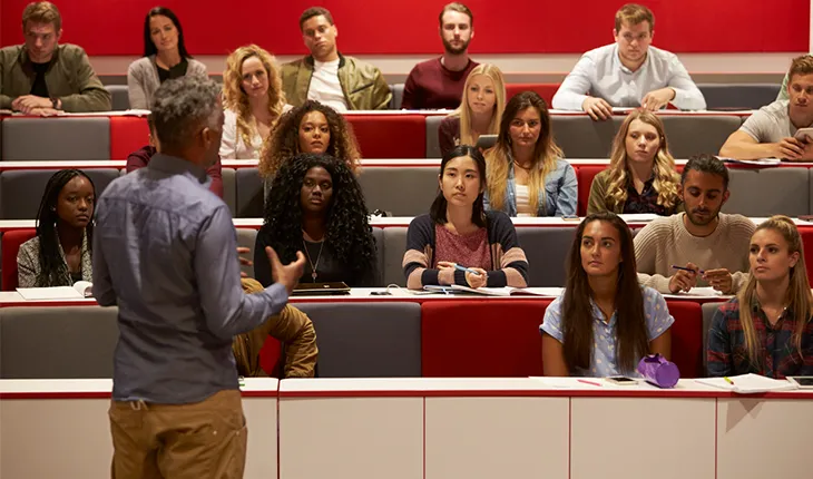 A lecturer presents to an engaged group of students in a lecture theatre with a red backdrop.
