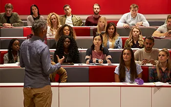 A lecturer presents to an engaged group of students in a lecture theatre with a red backdrop.