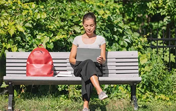 A student sits on a bench, with her red backpack next to her, typing into a laptop