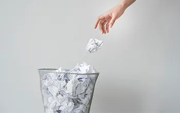 a hand throws a scrunched up piece of paper into a wastepaper basket, containing other scrunched up bits of paper