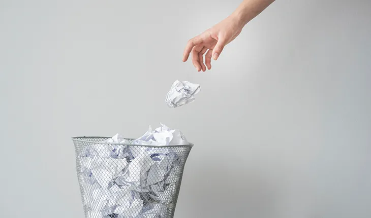 a hand throws a scrunched up piece of paper into a wastepaper basket, containing other scrunched up bits of paper