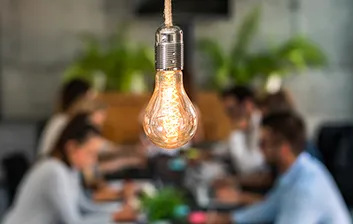 a group of facilities managers meet around a large table, a bright lightbulb above them signifies an idea