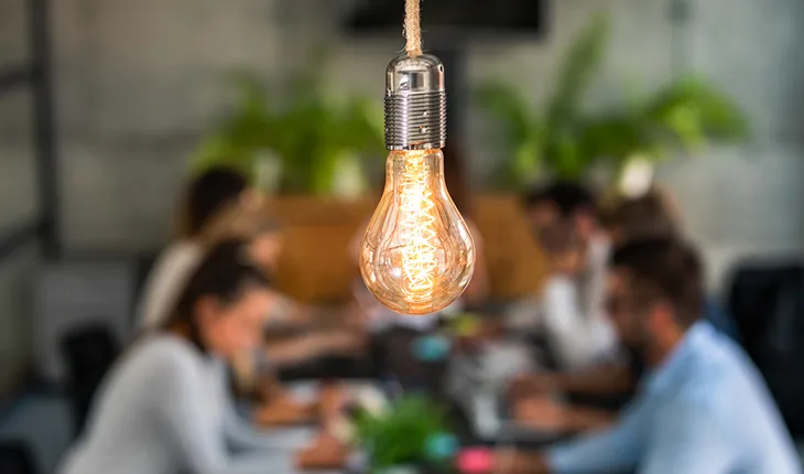 a group of facilities managers meet around a large table, a bright lightbulb above them signifies an idea