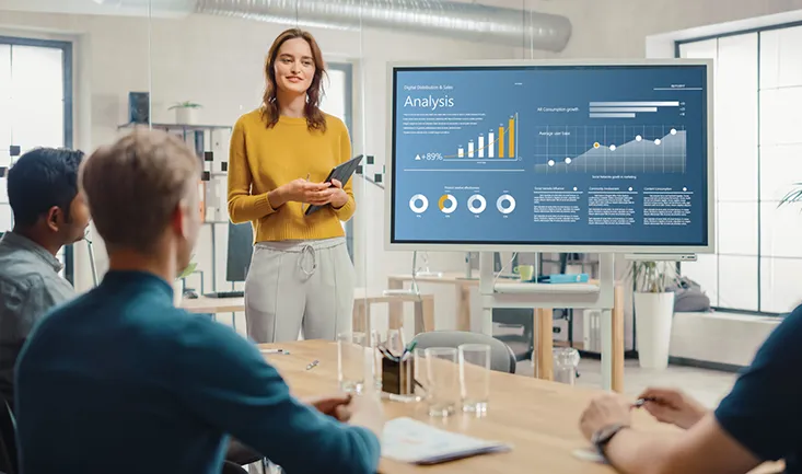 a young women presents graphically displayed data on a digital whiteboard to a boardroom full of people
