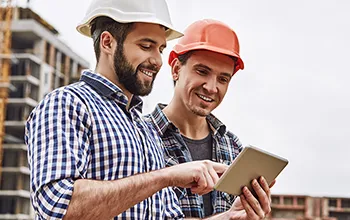 A facilities manager performs an induction for a tradesperson. Two caucausian men in hard hats look at information on an iPad.