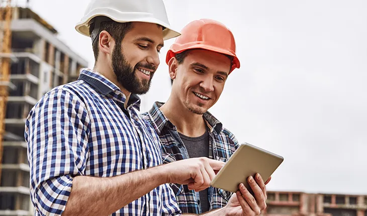 A facilities manager performs an induction for a tradesperson. Two caucausian men in hard hats look at information on an iPad.