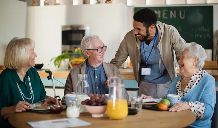 A male presenting aged care worker laughs with some of the residents who are sitting around a table having breakfast