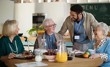 A male presenting aged care worker laughs with some of the residents who are sitting around a table having breakfast