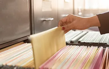 A manicured hand selects a manila folder out of a stuffed filing cabinet.