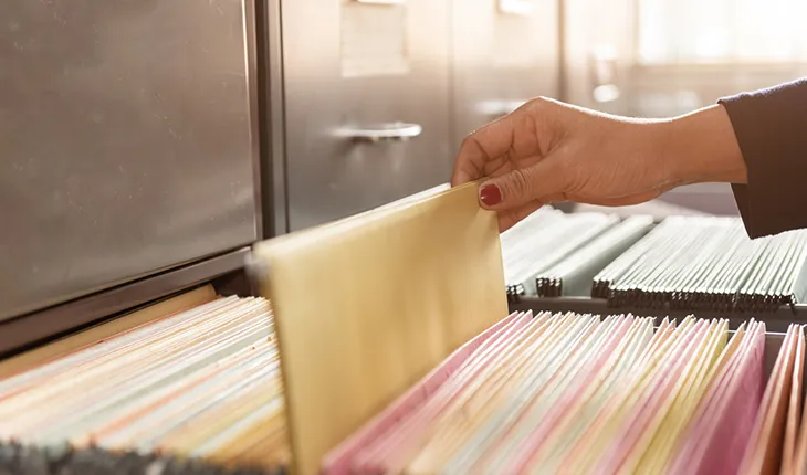 A manicured hand selects a manila folder out of a stuffed filing cabinet.