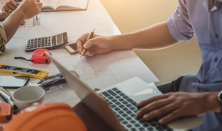 A facilities manager looks at a bunch of papers on their desk, which also has some maintenance tools and an orange hard hat on it. 