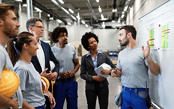 A male facilities manager speaks to a group of collegues while gesturing at a whiteboard with various information on it, the group are dressed to represent all different departments and levels in the organisational heirarchy