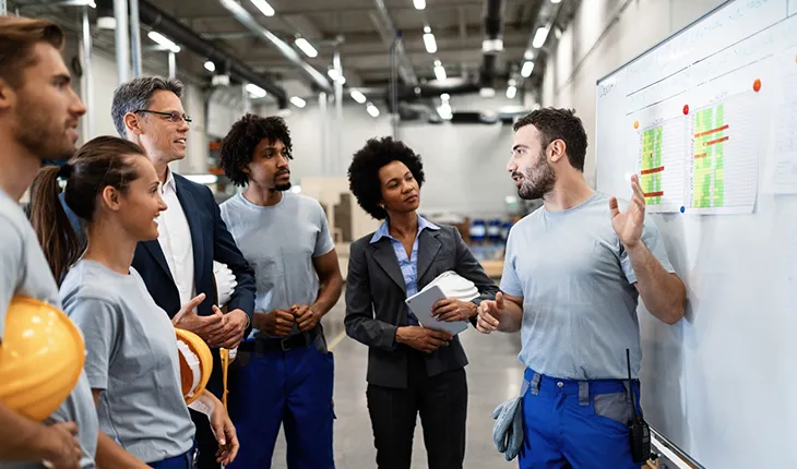 A male facilities manager speaks to a group of collegues while gesturing at a whiteboard with various information on it, the group are dressed to represent all different departments and levels in the organisational heirarchy