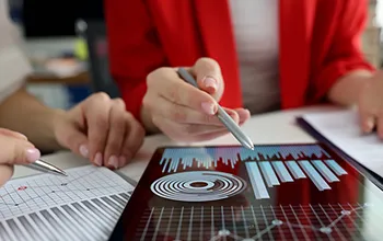 a woman in a red jacket uses a stylus to adjust graphs on an ipad while another collegue takes notes