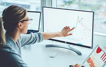 A young professional female follows a line graph on her monitor with her finger, dragging the graph to project potential scenarios.