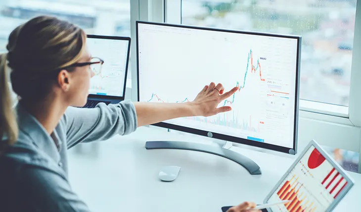 A young professional female follows a line graph on her monitor with her finger, dragging the graph to project potential scenarios.