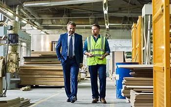 A man in a suit walks through a warehouse with a facilities manager wearing high vis safety gear