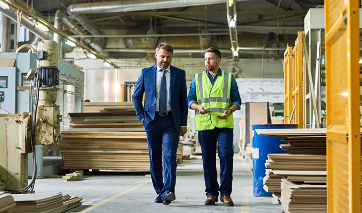 A man in a suit walks through a warehouse with a facilities manager wearing high vis safety gear