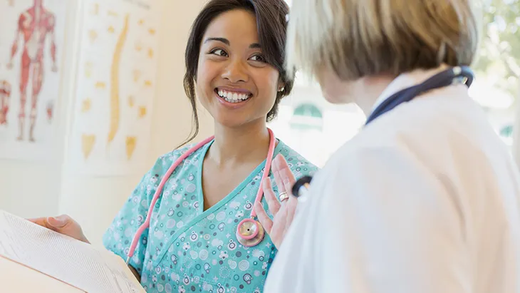 A young female-presenting nurse smiles at a doctor while they discuss a chart in a clinical setting