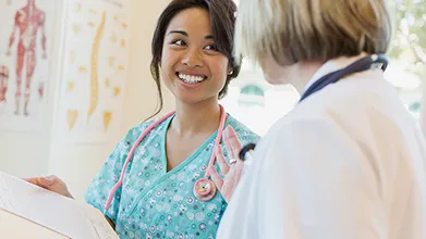 A young female-presenting nurse smiles at a doctor while they discuss a chart in a clinical setting