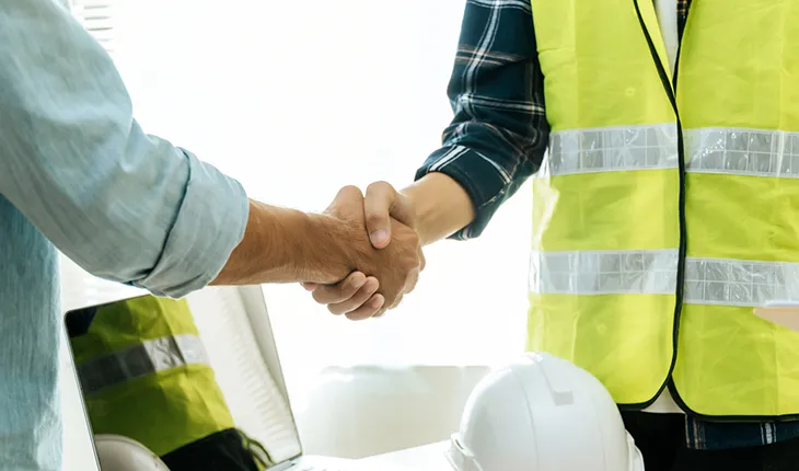 A facilities manager and a contractor wearing a high vis vest shake hands over a laptop